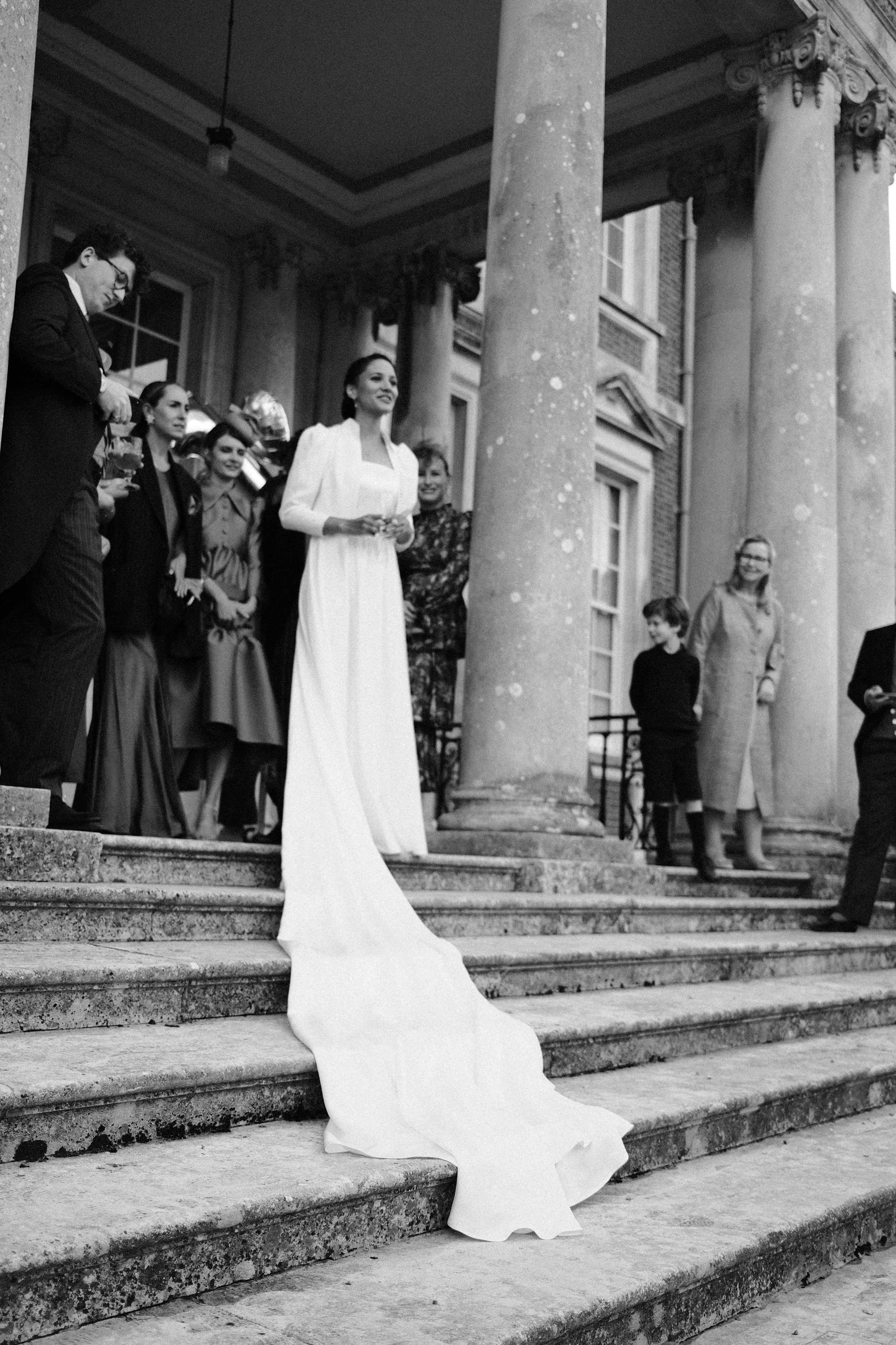 Eine Frau in einem langen weißen Kleid steht an den Stufen vor einem historischen Gebäude mit großen Säulen, umgeben von mehreren Menschen, darunter ein Kind und eine ältere Frau.