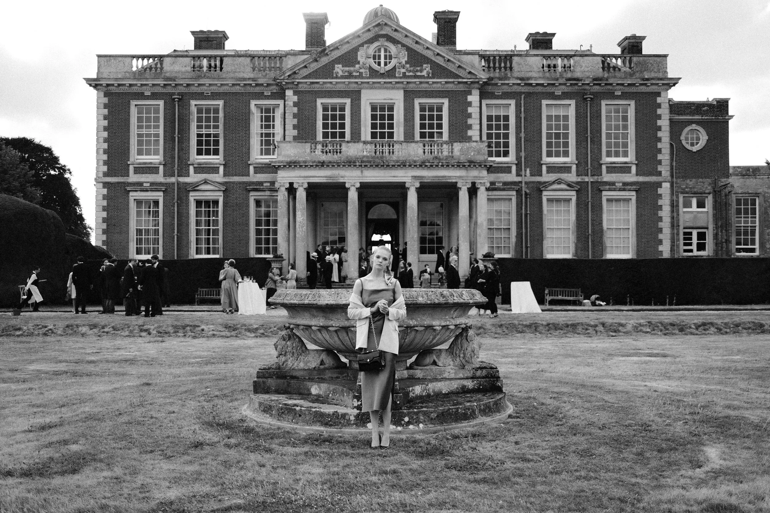 Schwarz-weiß-Foto eines großen Herrenhauses im klassischen Stil mit vielen Fenstern und Säulen, im Vordergrund eine junge Frau, die vor einem alten Brunnen steht, und im Hintergrund Menschen bei einer Veranstaltung auf der Rasenfläche.