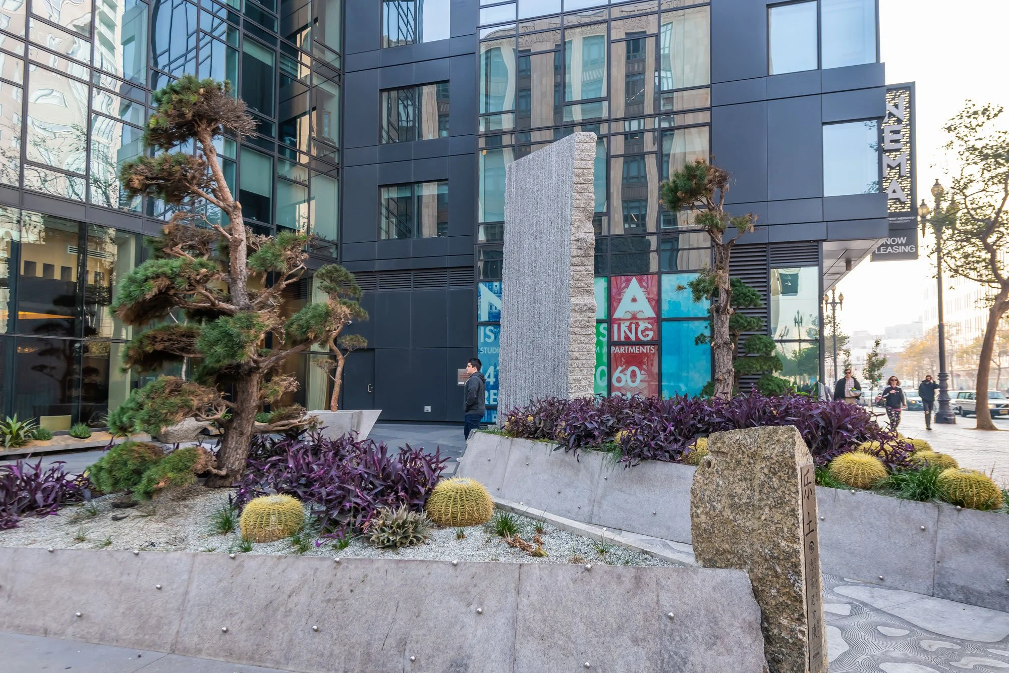 Modern city sidewalk scene with a small landscaped area featuring two twisted pine trees, purple-leaf plants, yellow cacti, and a large textured stone sculpture, with a black glass building and pedestrians in the background.