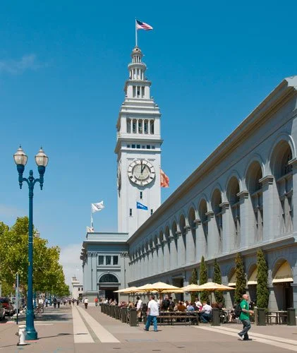 Ferry Building with its clock tower and flags in San Francisco, California, with people dining outside under umbrellas on a sunny day.