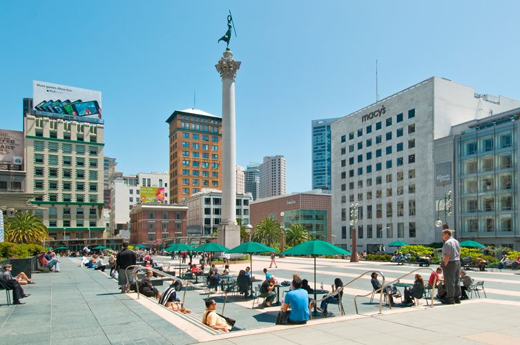 City square with people sitting on benches, green umbrellas, and a tall monument with a statue on top, surrounded by modern high-rise buildings.