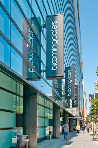 Exterior view of Bloomingdale's department store with blue glass facade and vertical store signs, with shoppers walking on the sidewalk.