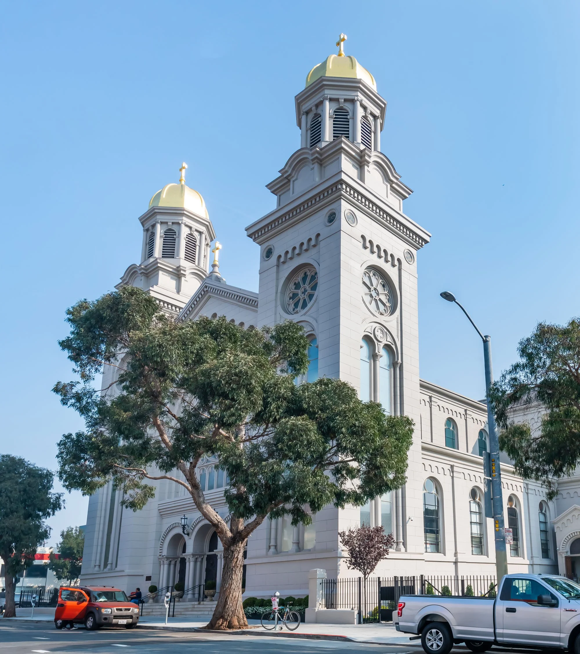 A white church with two tall towers topped with gold domes and crosses, surrounded by trees, parked cars, and a bicycle on a city street with a clear blue sky.
