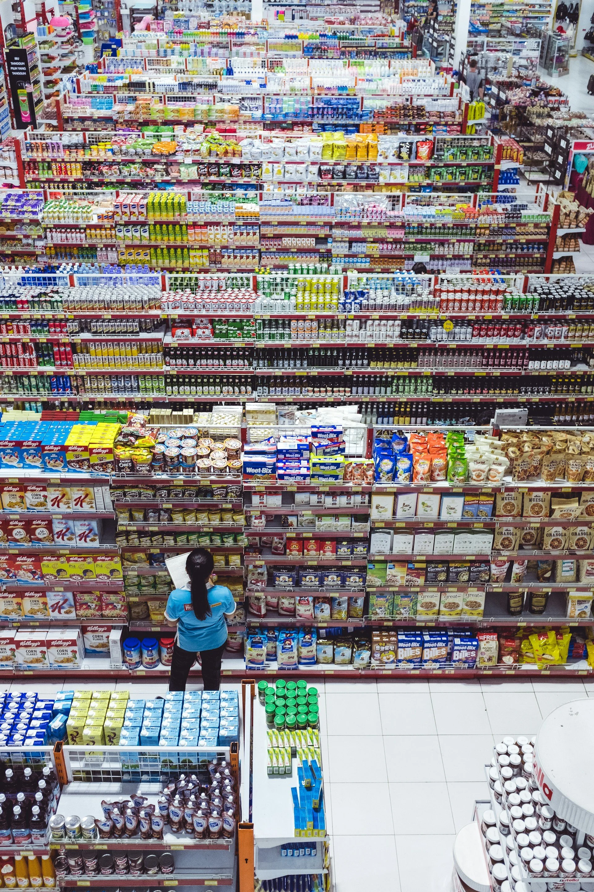 Blick auf die Regale in einem Supermarkt, gefüllt mit verschiedenen verpackten Lebensmitteln und Getränken. Eine Verkäuferin steht vor einem Regal und durchstöbert Produkte.