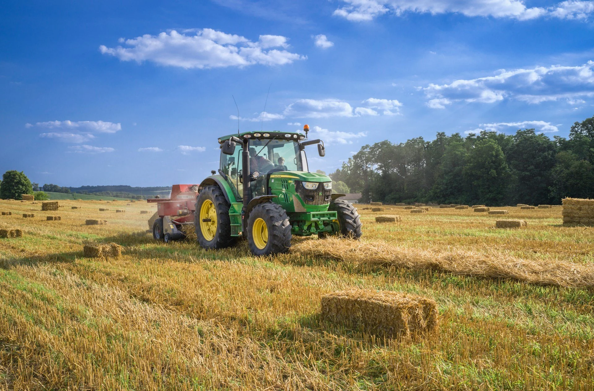 Traktor auf einem Feld bei Sonnenlicht, umgeben von Heuballen und Bäumen.