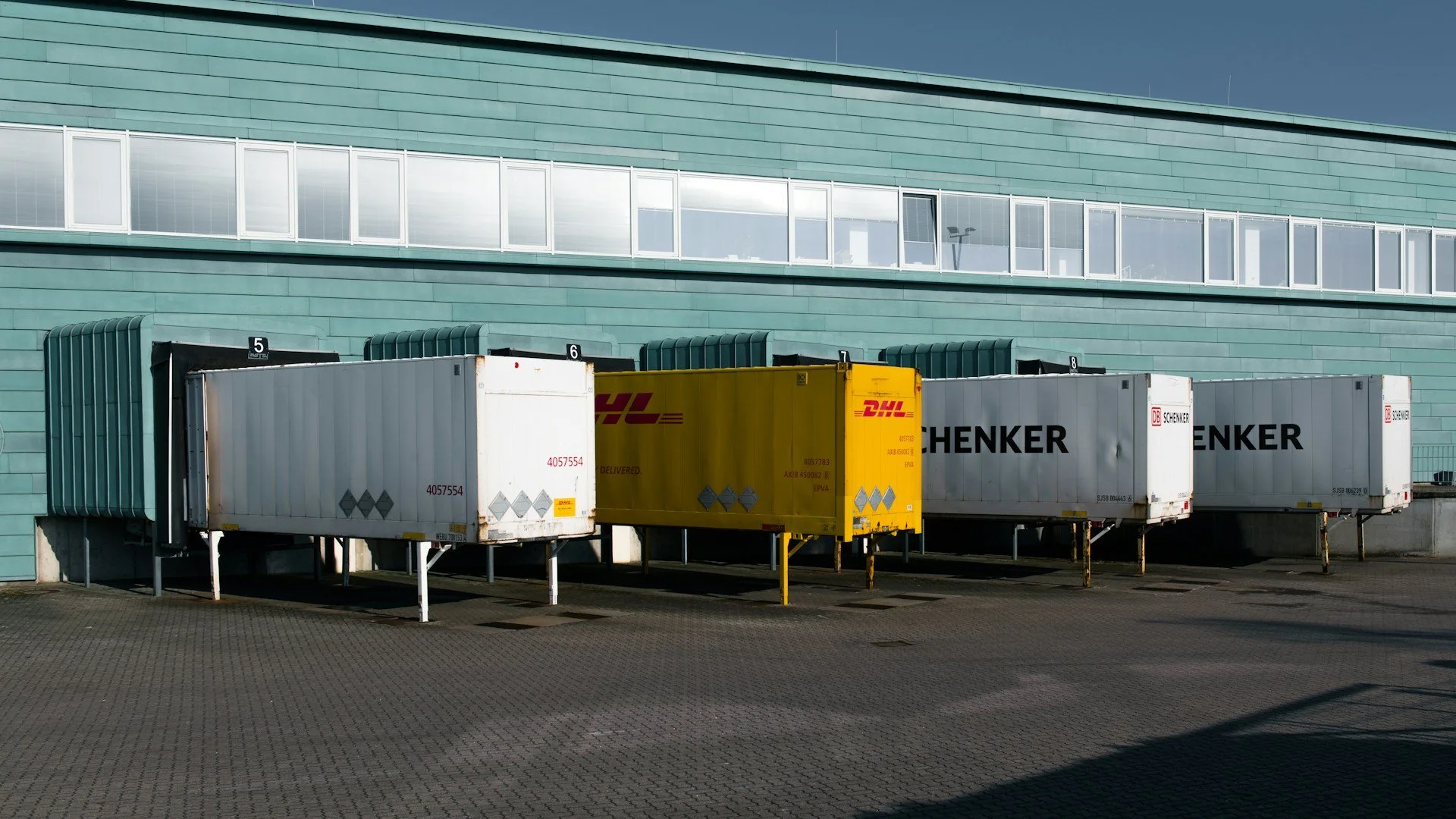 Mehrere Lieferwagen und Container vor einem modernen, blauen Gebäudekomplex, die auf einer abgezäunten Fläche stehen.