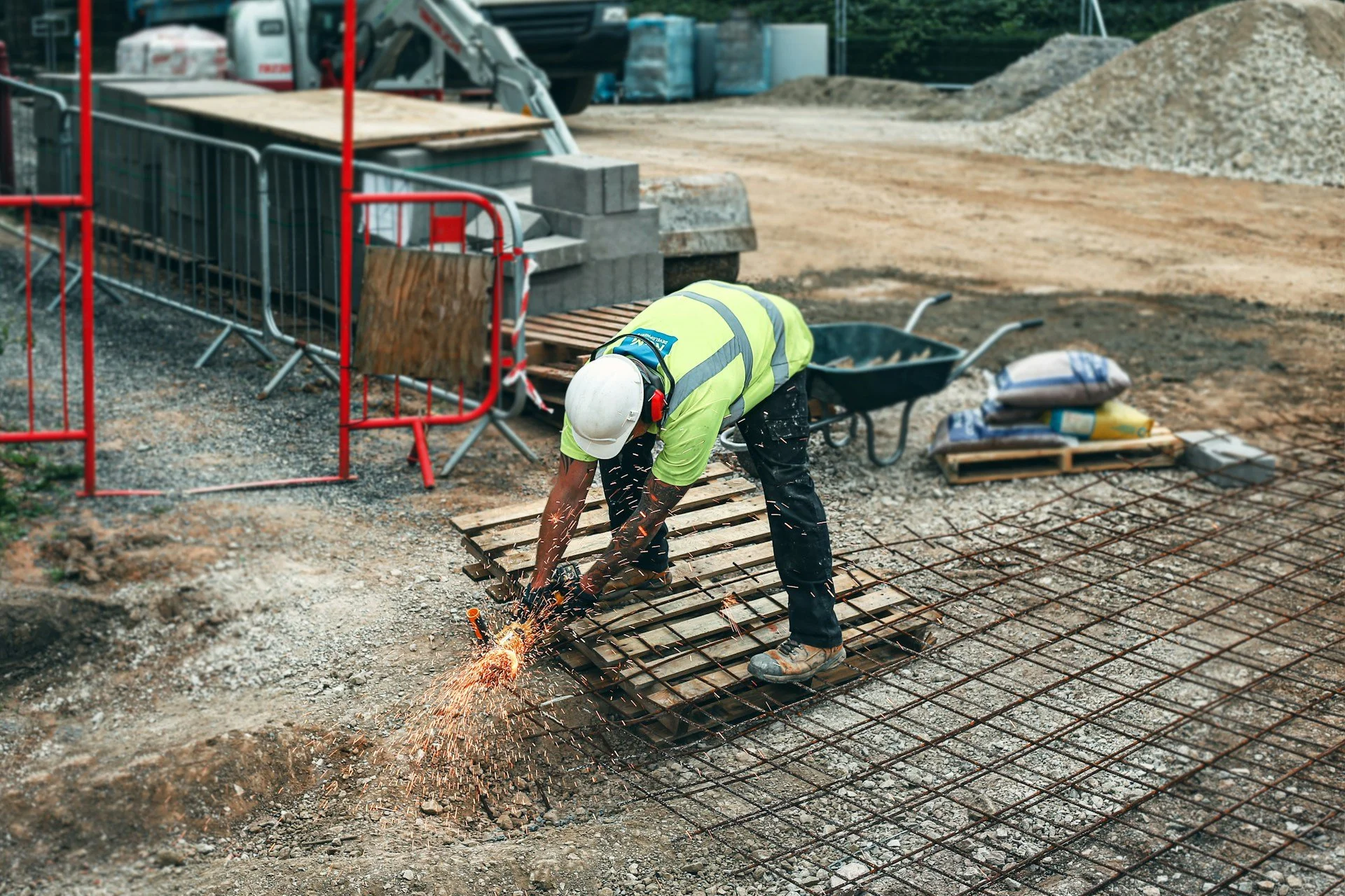 Arbeiter auf einer Baustelle schweißt Stahlbewehrung, trägt Schutzhelm und Sicherheitsweste, umgeben von Baumaterialien und Maschinen.