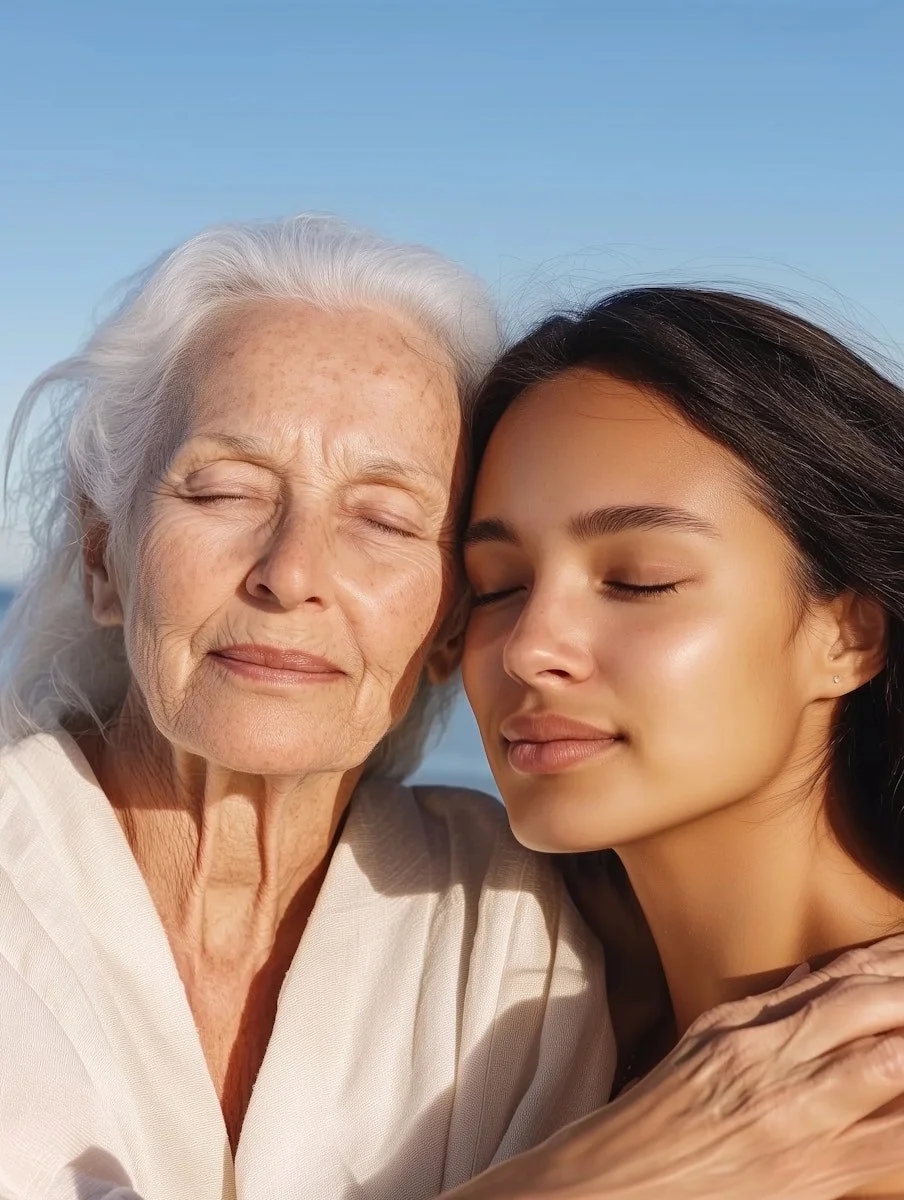 A young woman and an elderly woman are close together with their eyes closed, smiling softly, enjoying a peaceful moment outside with a blue sky background.