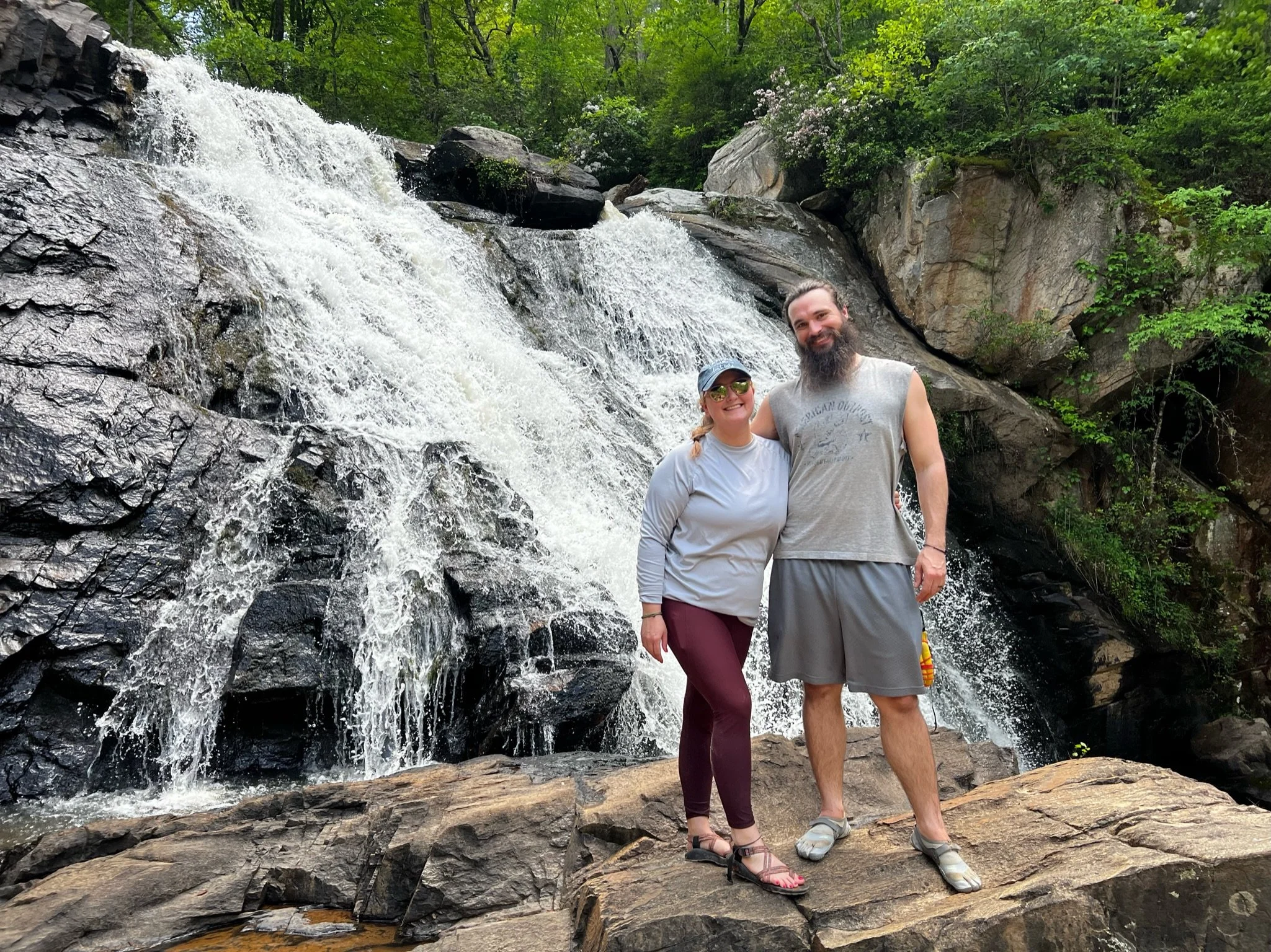 A man and woman smiling and posing by a waterfall in a forest.