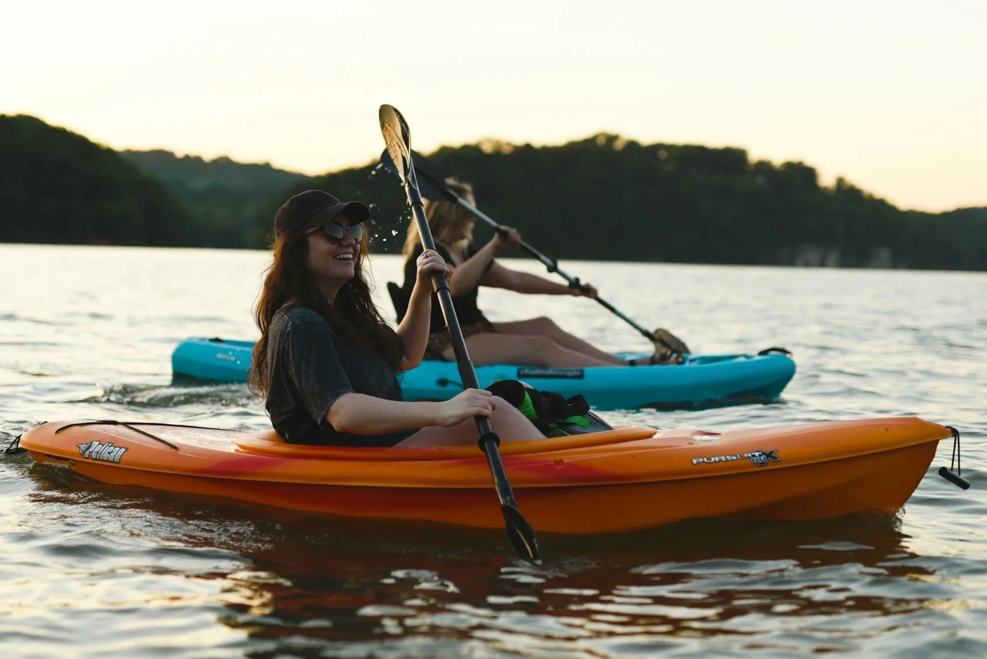 Two women kayaking on a lake during sunset, smiling and enjoying the outdoors with mountains and trees in the background.