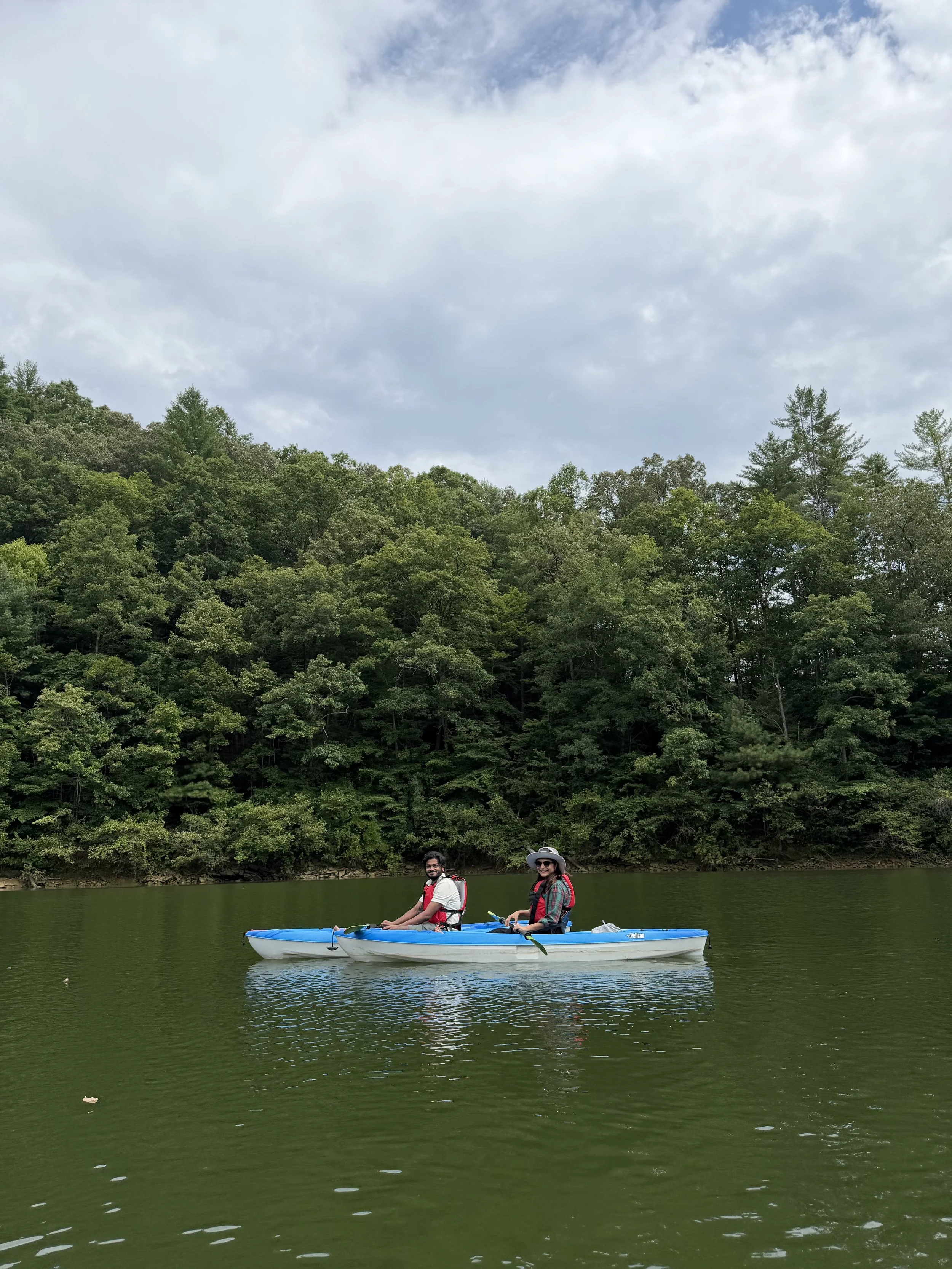 Two people kayaking on a calm river with lush green trees on a forested hillside behind them under a partly cloudy sky.