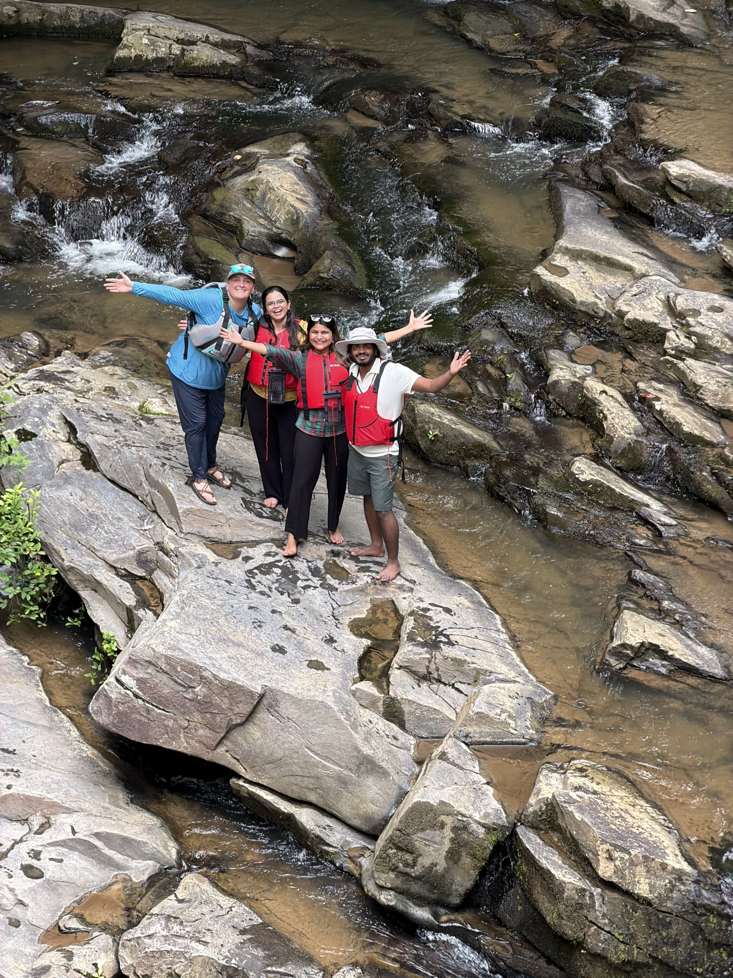 Four people standing on rocks by a flowing stream, smiling and spreading their arms, surrounded by natural rocky landscape.