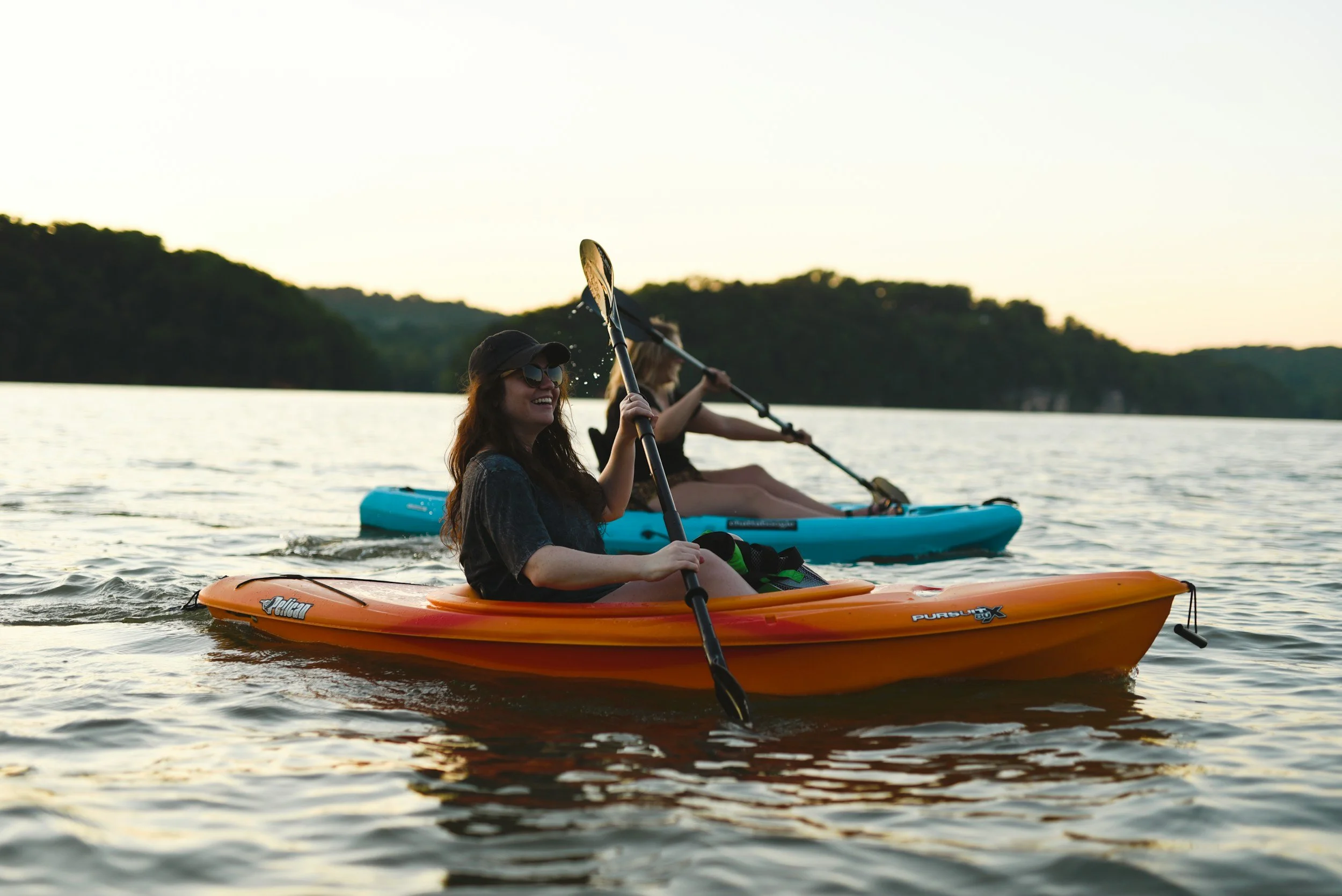 Two women kayaking on a lake at sunset, one in an orange kayak and the other in a blue kayak, smiling and enjoying the outdoors.