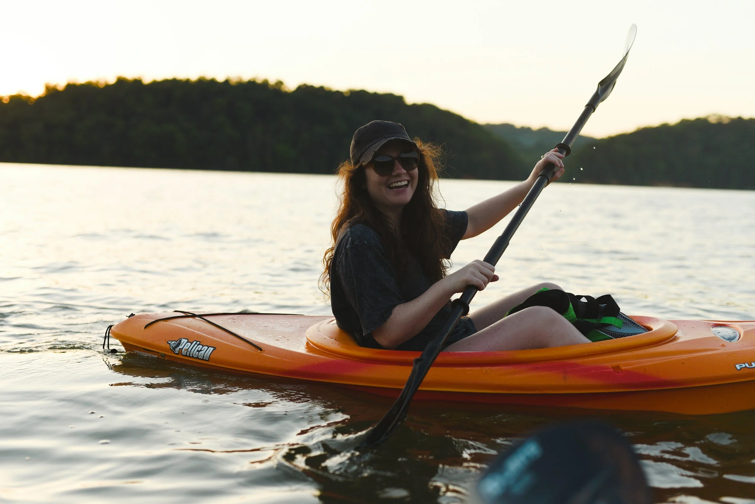 A woman with curly hair, sunglasses, and a hat happily kayaking on a lake at sunset, holding a paddle and wearing a black t-shirt.