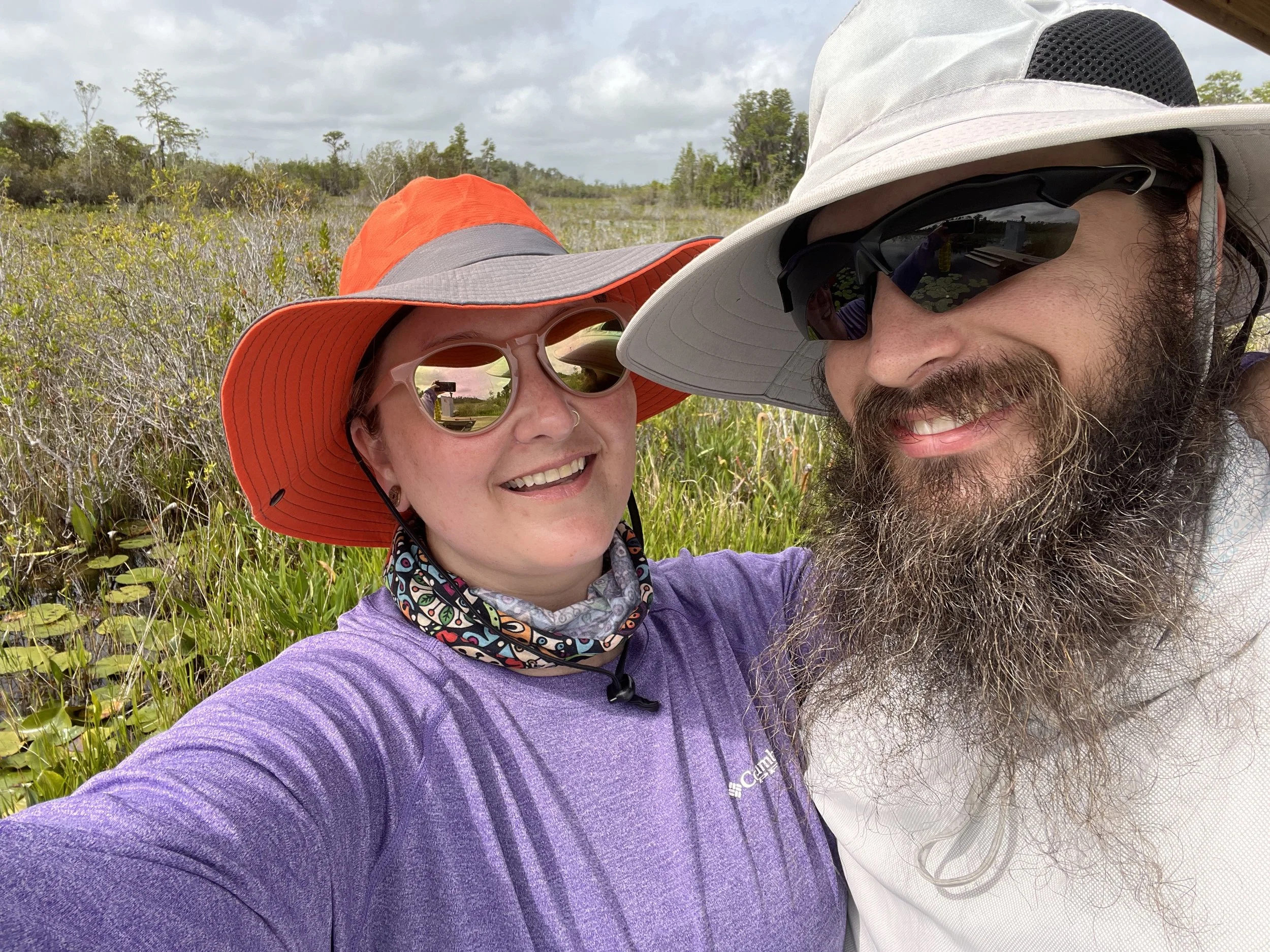 Two people outdoors wearing hats and sunglasses, smiling, with a marsh and trees in the background.