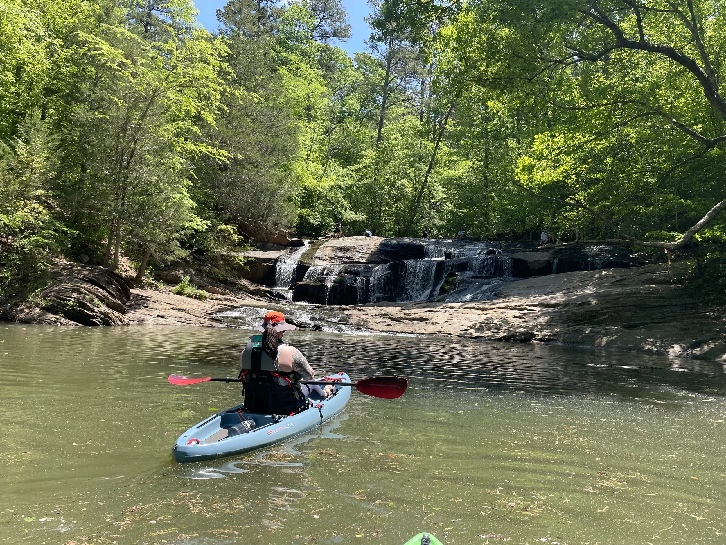 Person kayaking on a calm river with a waterfall and lush green trees in the background.