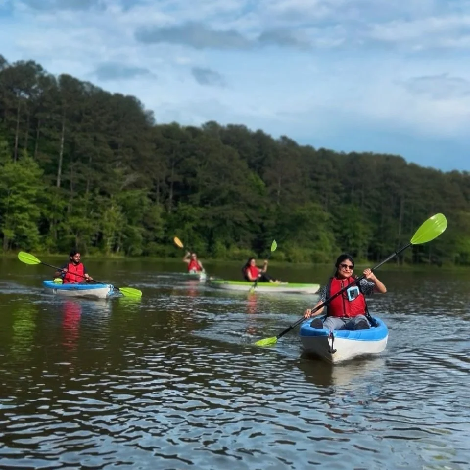 Four people kayaking on a lake with a forested shoreline, wearing life vests and paddling with green paddles.