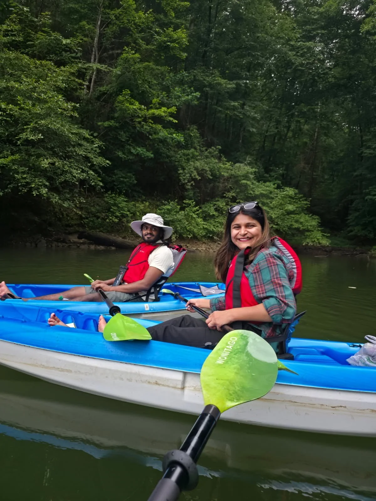 Two people kayaking on a calm river surrounded by lush green trees, smiling and holding paddles. The woman in the foreground is wearing a plaid shirt and sunglasses, while the man in the background is wearing a wide-brimmed hat and a white shirt.