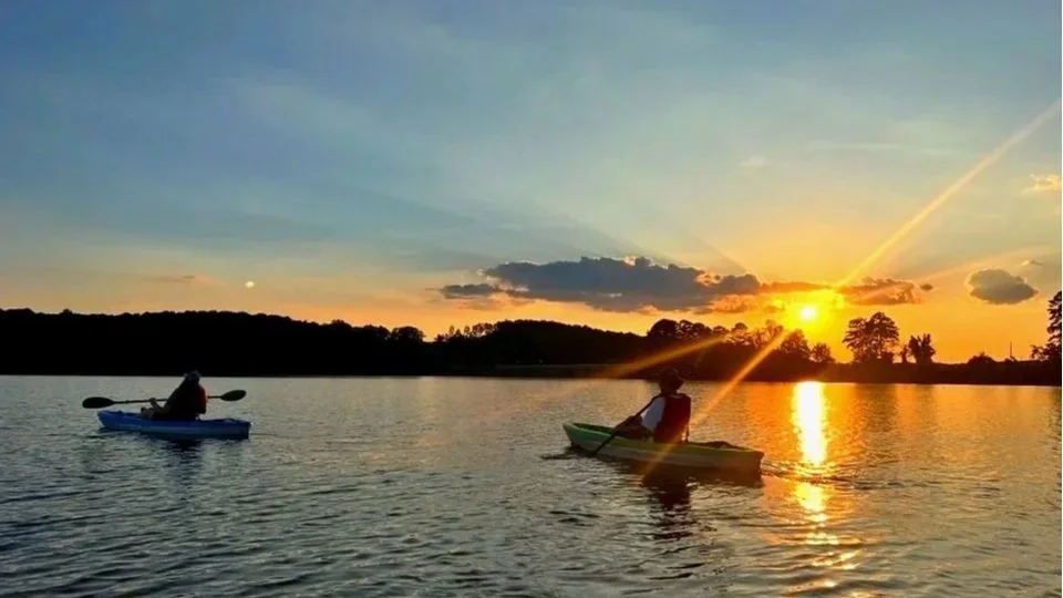 Two people kayaking on a lake at sunset with the sky painted in shades of orange, yellow, and blue, and trees silhouetted in the background.