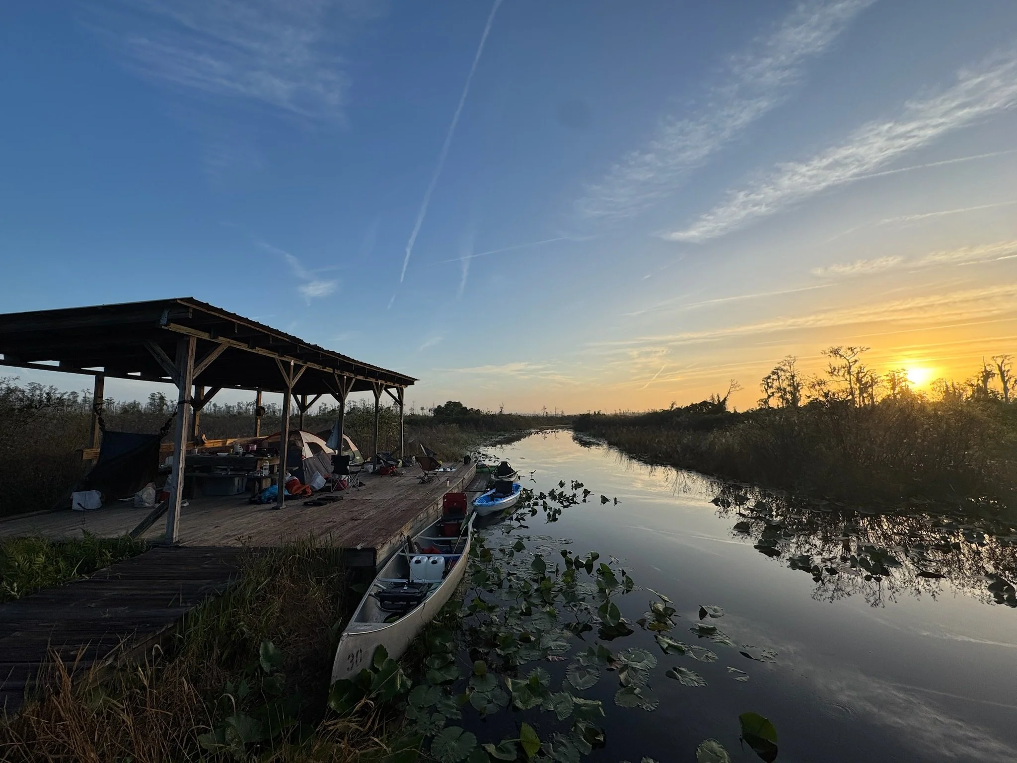 Sunset over a calm waterway with a wooden dock, boats, camping tents and a shelter, surrounded by trees and water lilies.