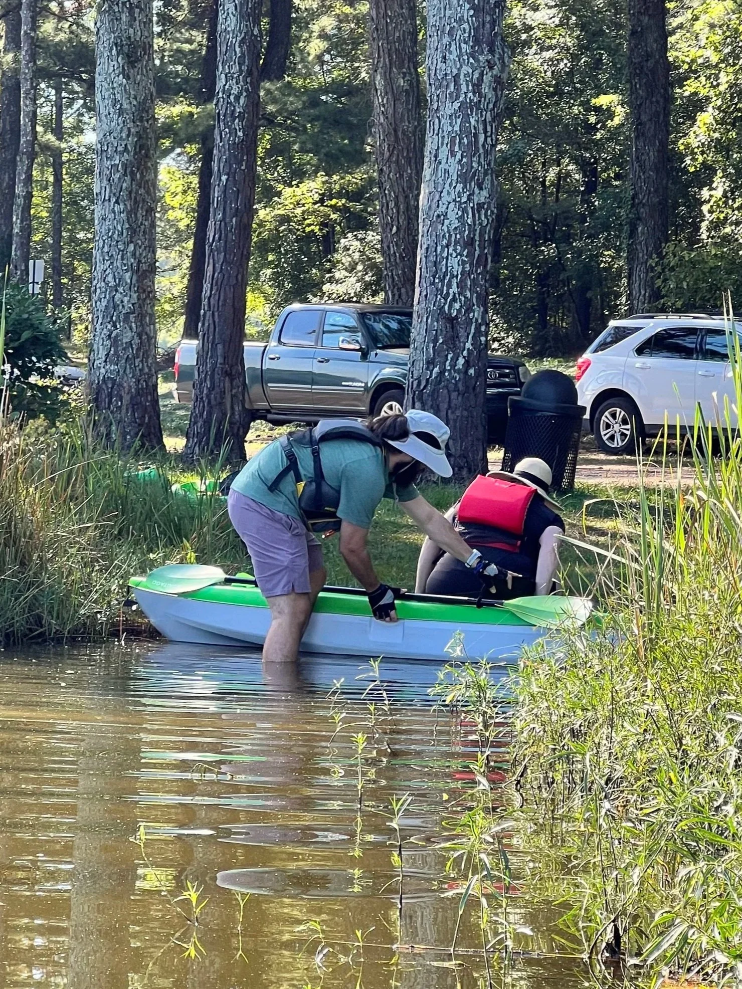 A person in a gray hat and blue shirt helping another person in a black hat and red life jacket into a green and white kayak in a river, with trees and parked cars in the background.