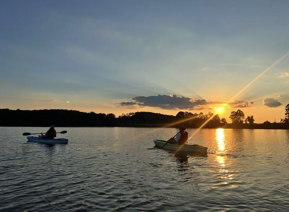 Two kayakers paddling on a calm lake during sunset, with silhouettes of trees and clouds in the background.