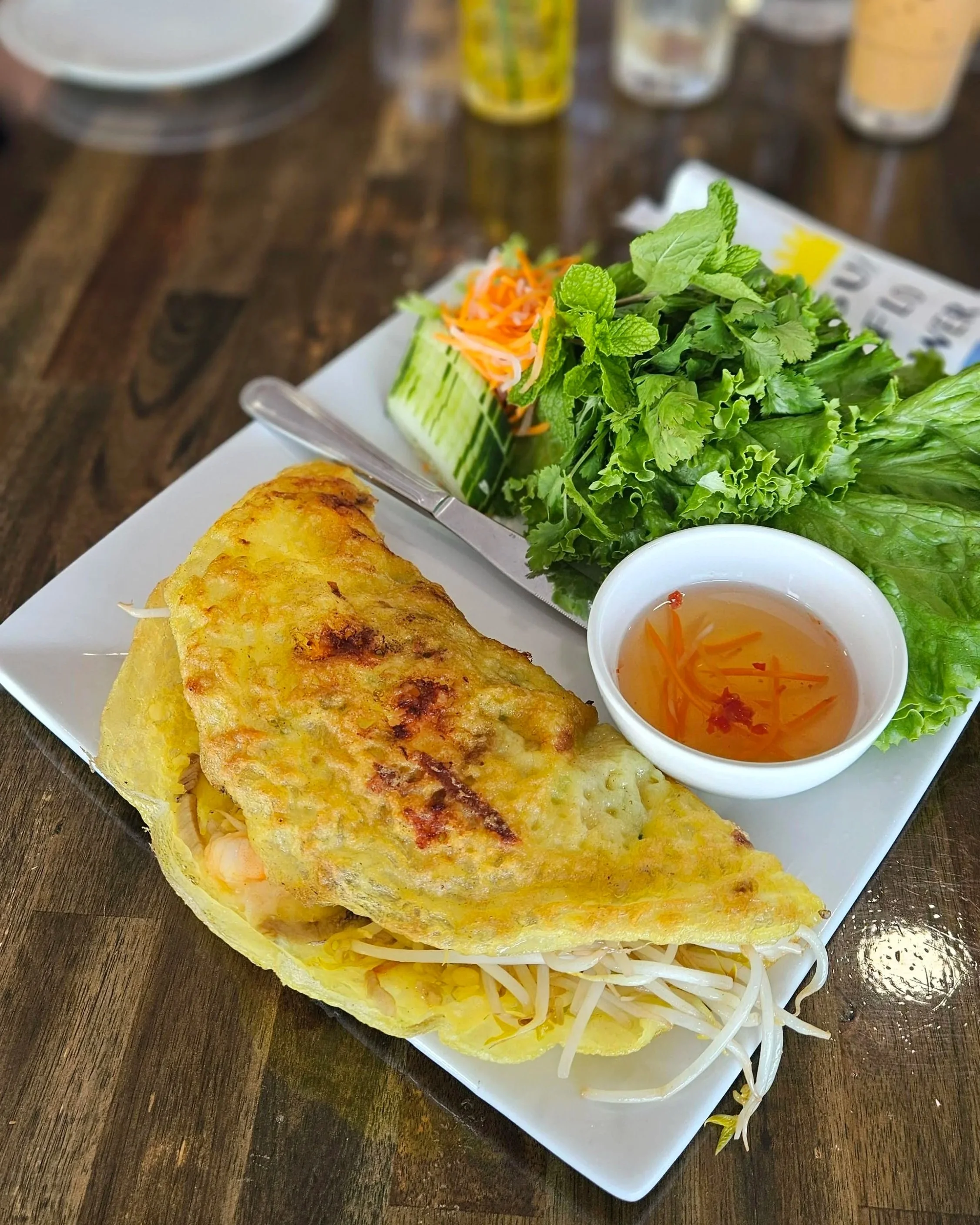 square plate with yellow crispy vietnamese crepe with fresh shrimp and bean spouts spilling out of the opening accompanied with a dipping sauce and fresh vegetables.