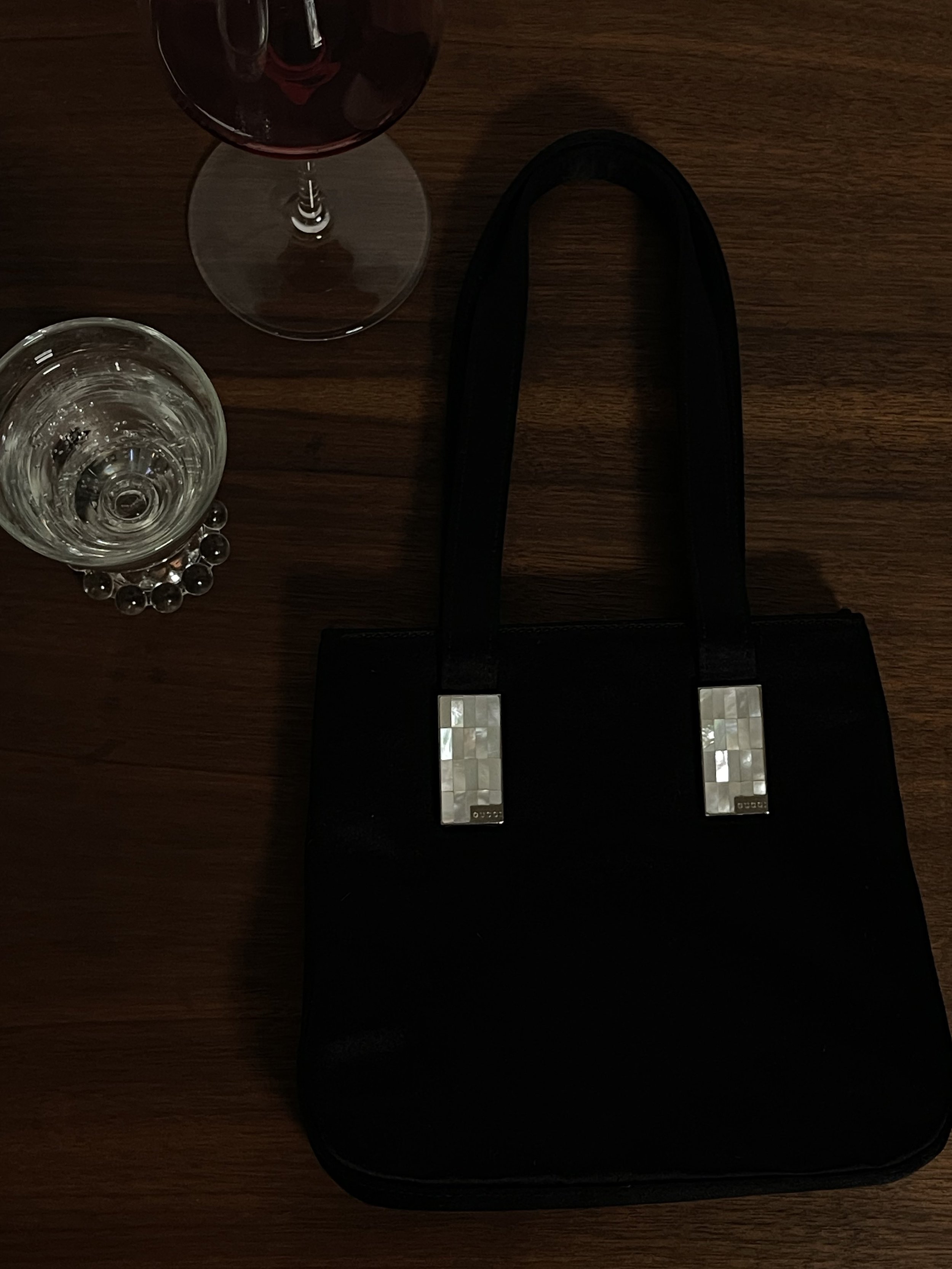 A black Tom Ford Gucci silk evening bag with mother-of-pearl accents on the handles, placed on a wooden table, alongside a glass of red wine and a glass of clear water.