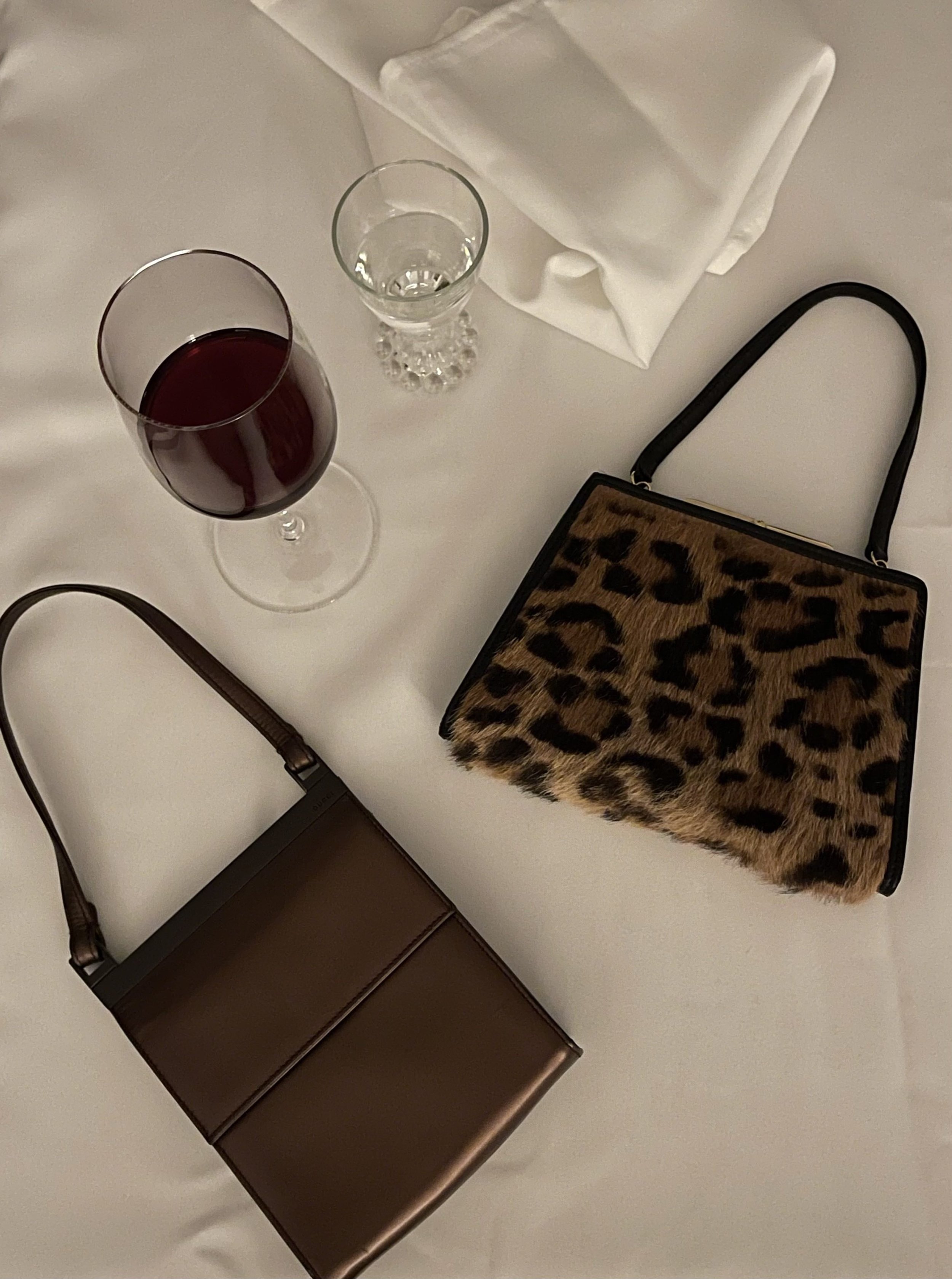 A table setting with two handbags a leopard Loewe evening bag and a brown Tom Ford Gucci evening bag, a glass of red wine, a glass of water, and a folded white napkin.