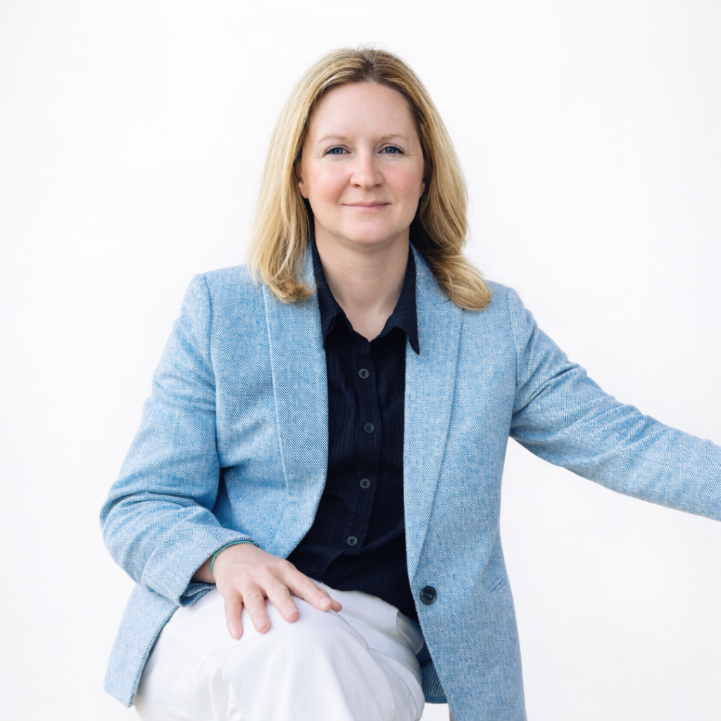 Woman with blonde hair wearing a blue blazer and black shirt sitting against a white background.