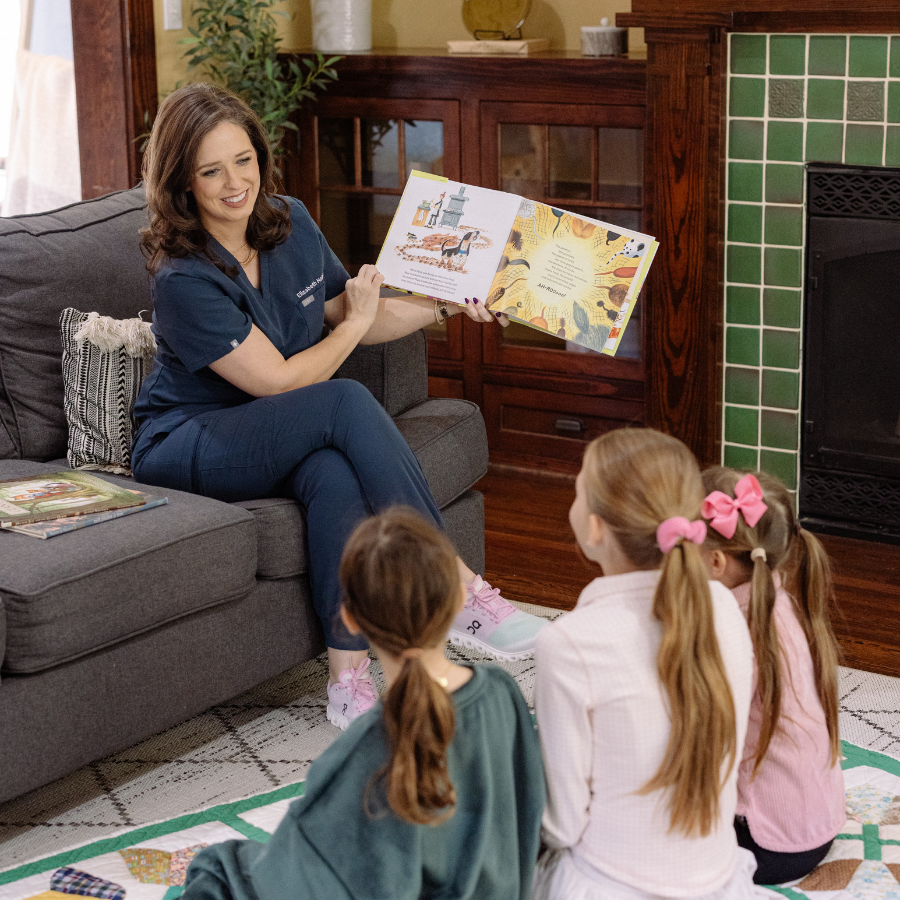 A pediatric doctor reading a colorful picture book to three young girls with ponytails and pink bows, sitting on a patterned rug near a fireplace with green tiles in a cozy living room.