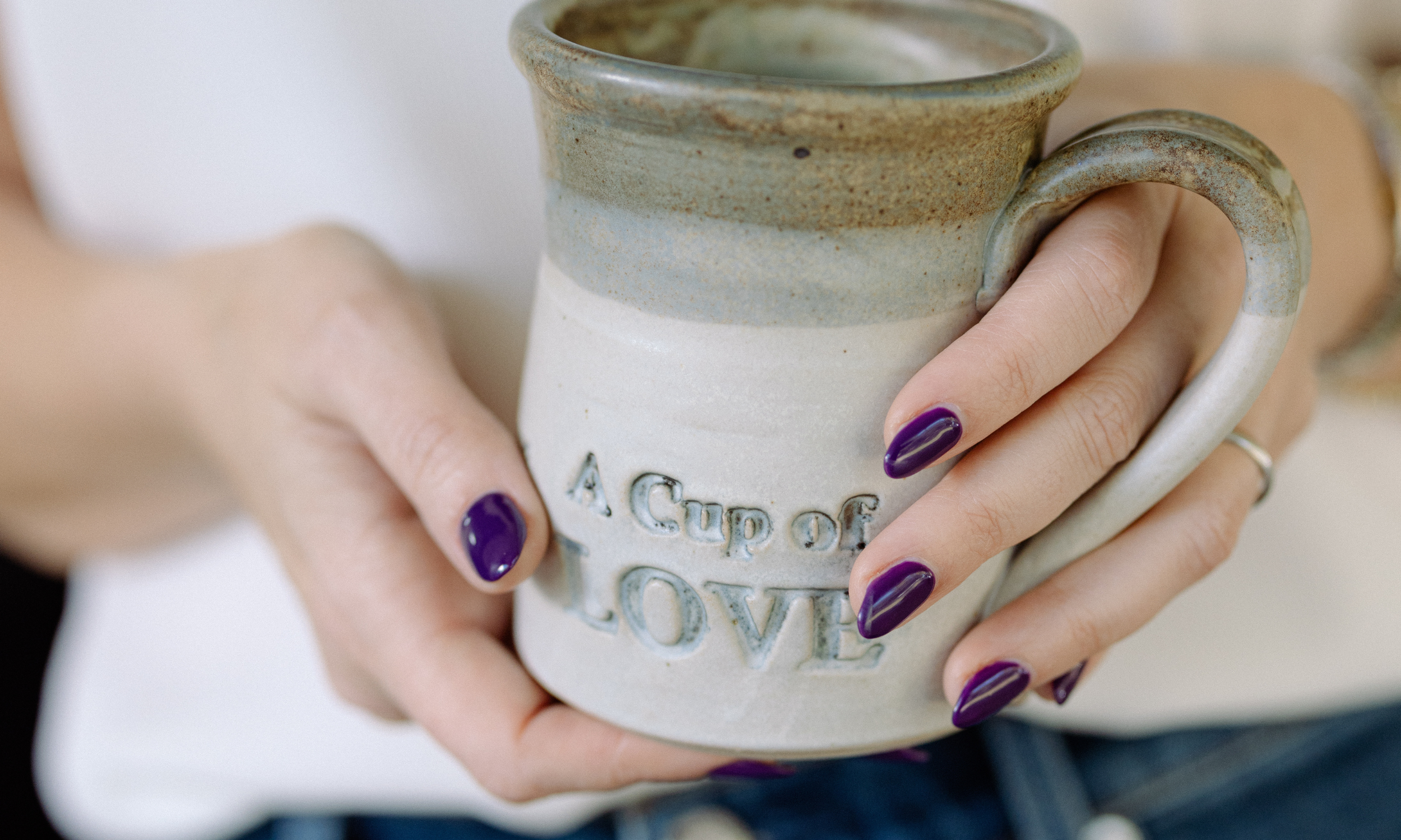 A person with purple nail polish holding a ceramic mug that has the words "A Cup of Love" embossed on it.