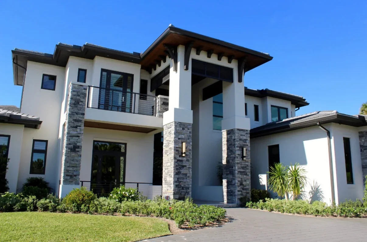 Modern two-story house with stone accents, large windows, and a front yard with green grass and plants under a clear blue sky.