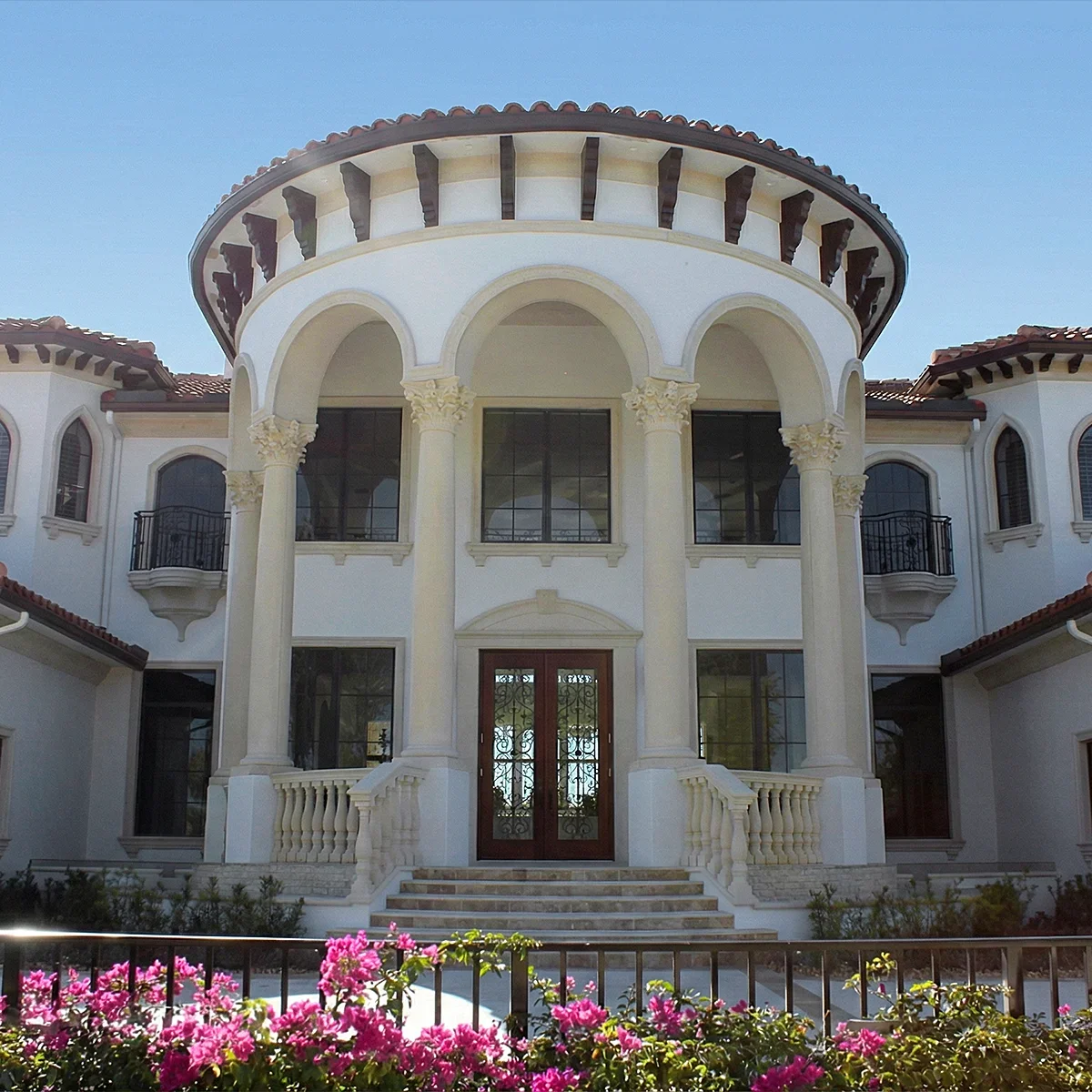 Front view of a large, elegant mansion with colonial architectural style, featuring tall columns, arched windows, and a balcony, with pink flowers in the foreground and a clear blue sky.