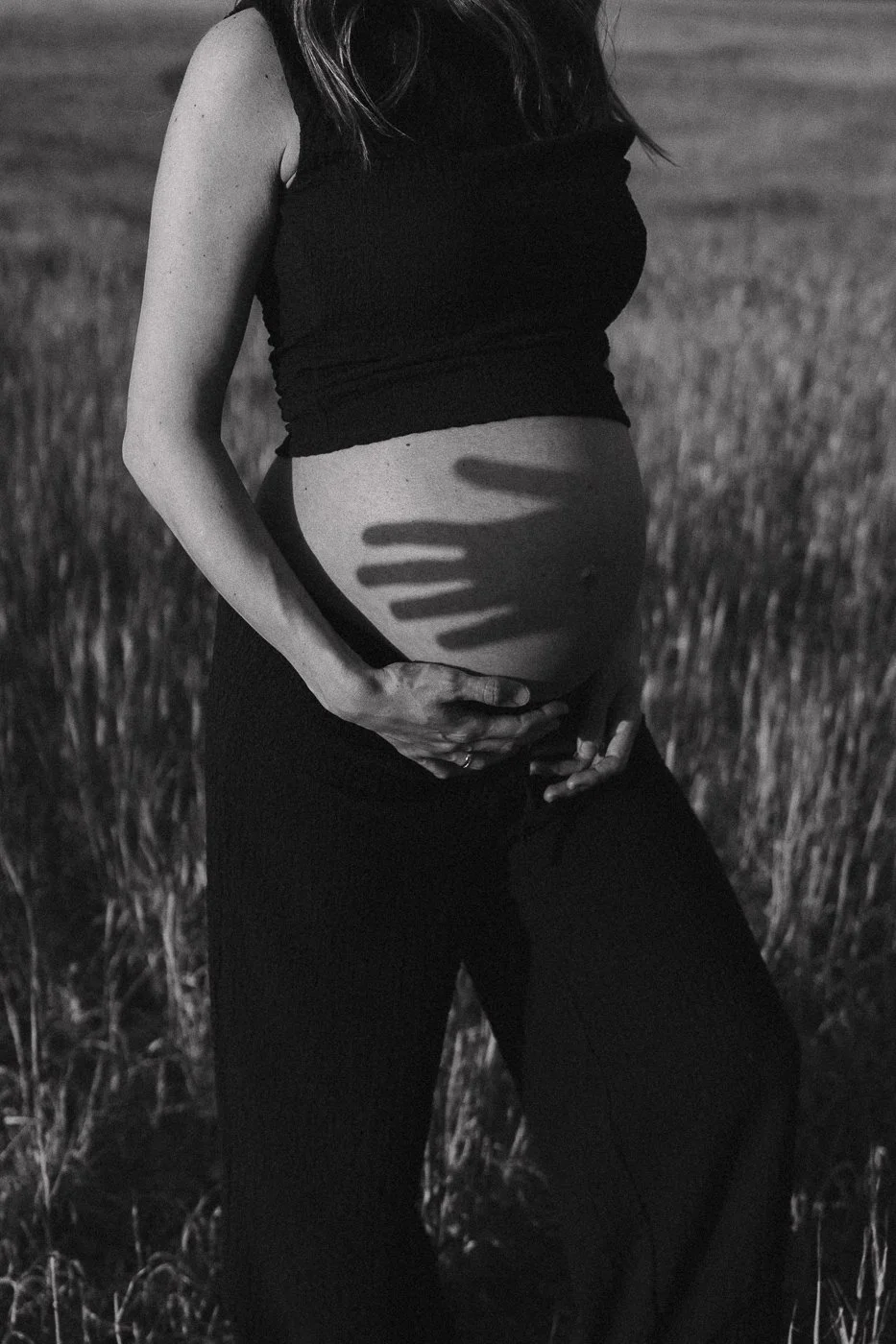Mujer embarazada en un campo, tocando su vientre y una sombra de mano en su abdomen, en blanco y negro.