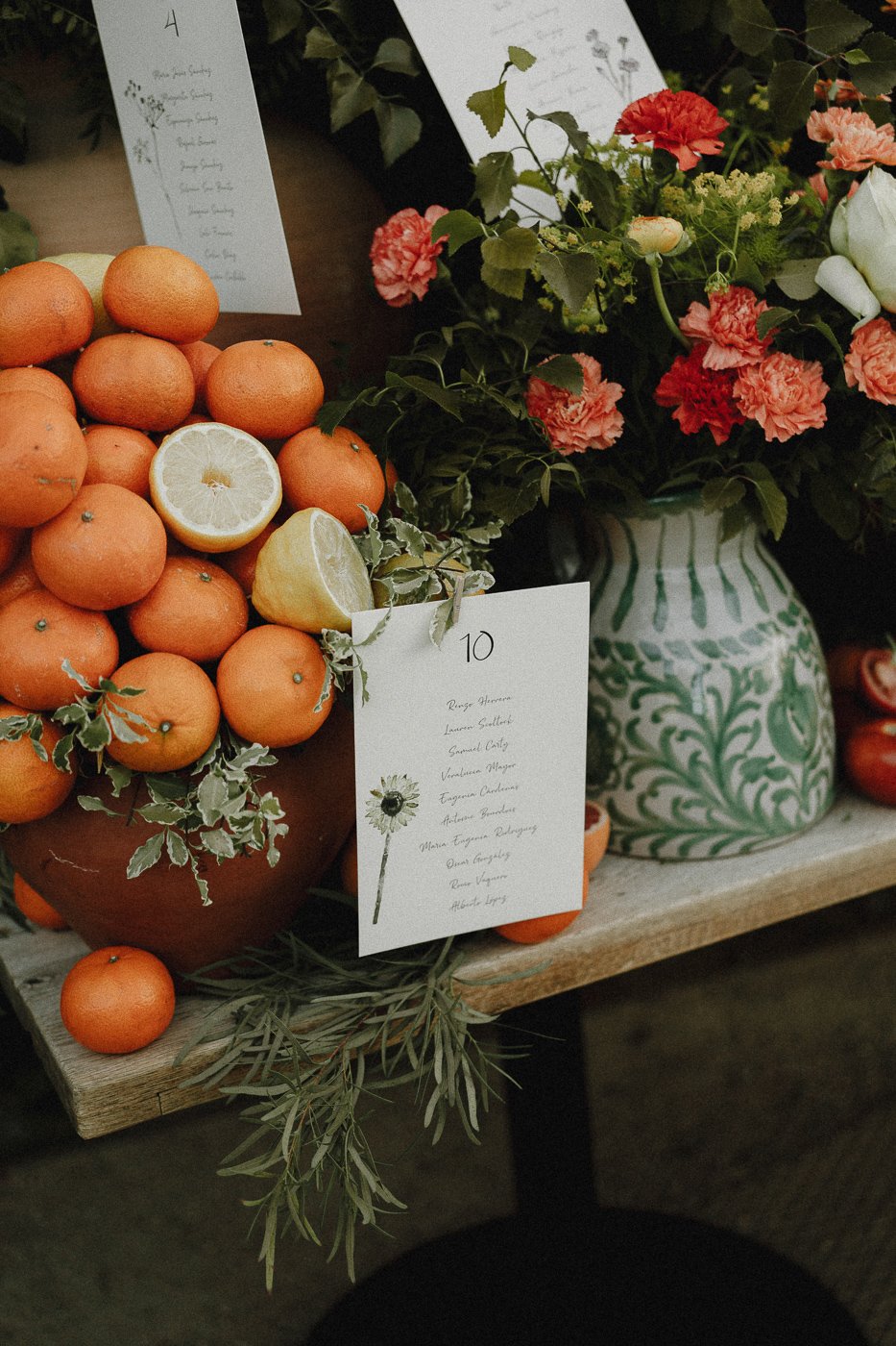 Un arreglo de naranjas y limones, con un ramo de flores rosas y verdes en un florero, en una mesa de madera. También hay una tarjeta con la lista de nombres, decorada con una ilustración de flor.