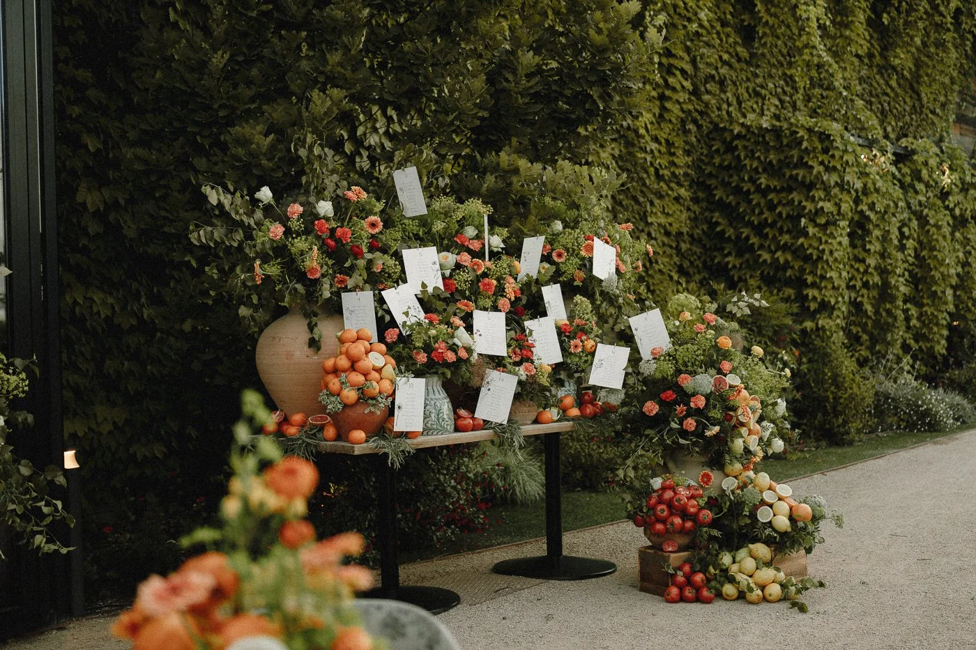 Decoración con flores y frutas en una mesa al aire libre con etiquetas colgantes, en un entorno con paredes cubiertas de hiedra.