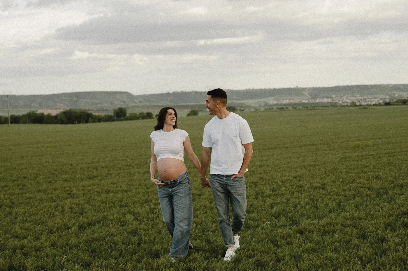 Pareja caminando en un campo verde, la mujer está embarazada y ambos se miran y sonríen, el cielo está nublado.