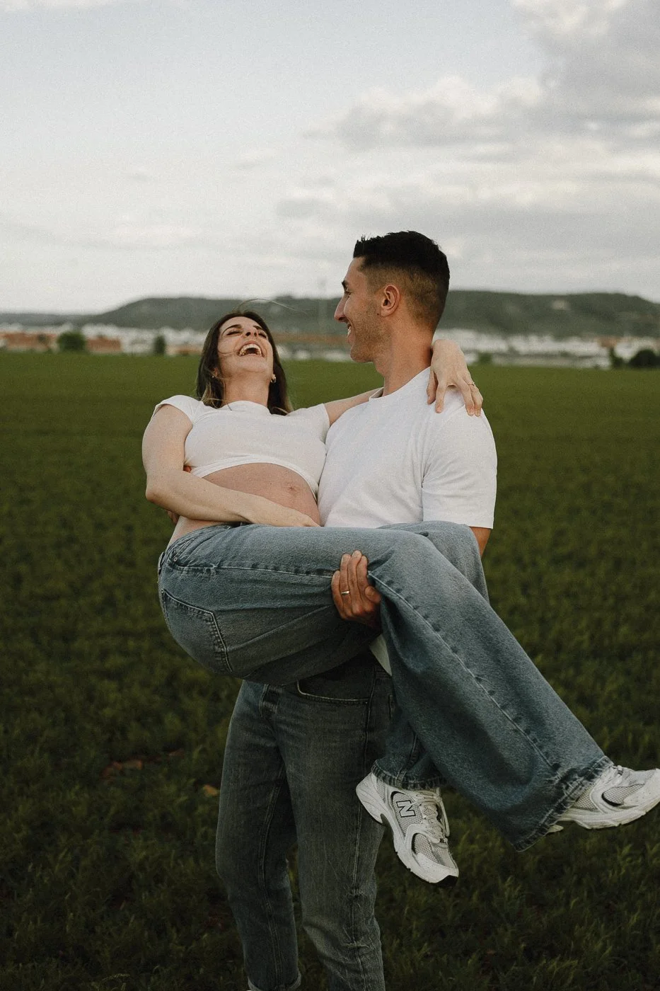 Una pareja feliz en un campo verde, el hombre sostiene a la mujer en brazos, ambos riendo y disfrutando del momento, paisaje de colinas y cielo nublado de fondo.
