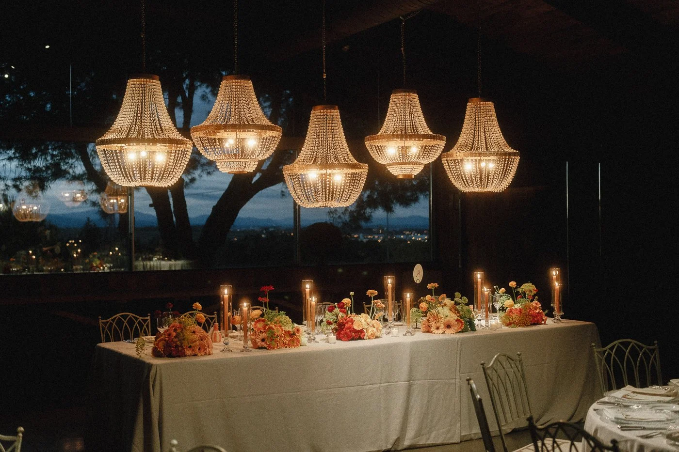 Mesa decorada con flores y velas, iluminada por candelabros de cristal y colgantes de lámparas en un ambiente nocturno con vista a la naturaleza.
