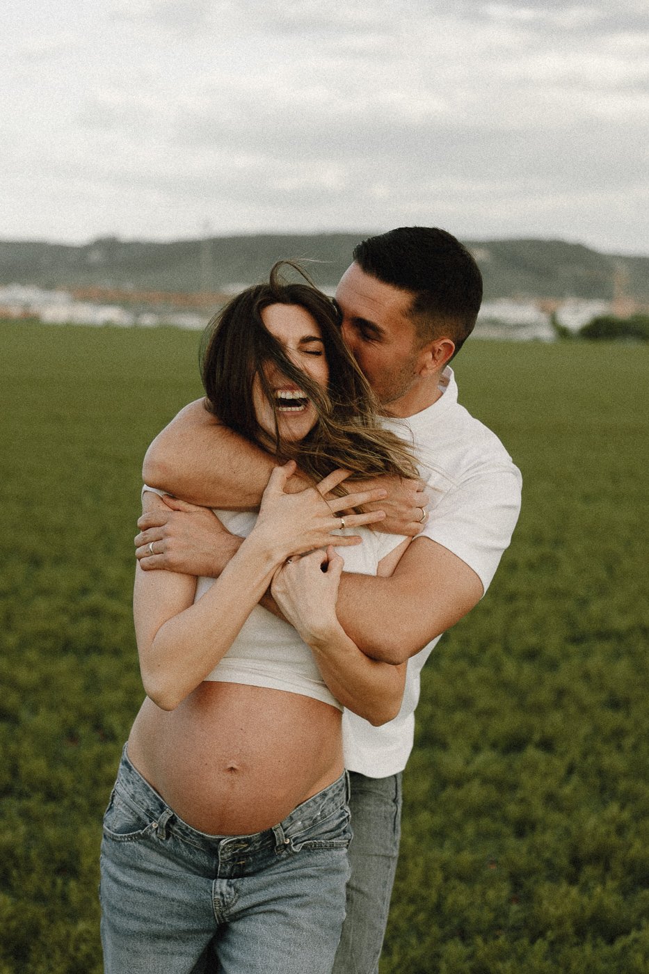 Pareja feliz en un campo, la mujer embarazada sonriendo y el hombre abrazándola.
