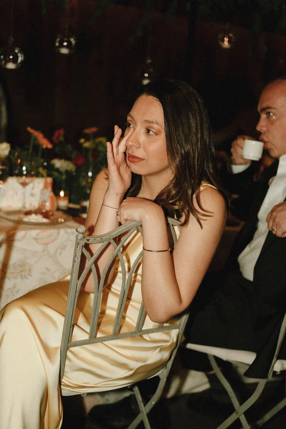 Una mujer con cabello castaño oscuro y piel clara, vestida con un vestido de color dorado, se encuentra sentada en una silla en un evento, con una expresión pensativa, tocándose la cara, en un entorno con decoración floral y tenue iluminación.