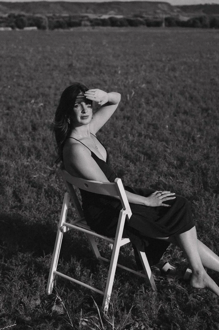 Mujer sentada en una silla en un campo abierto durante el atardecer, con la mano en la frente, en una fotografía en blanco y negro.
