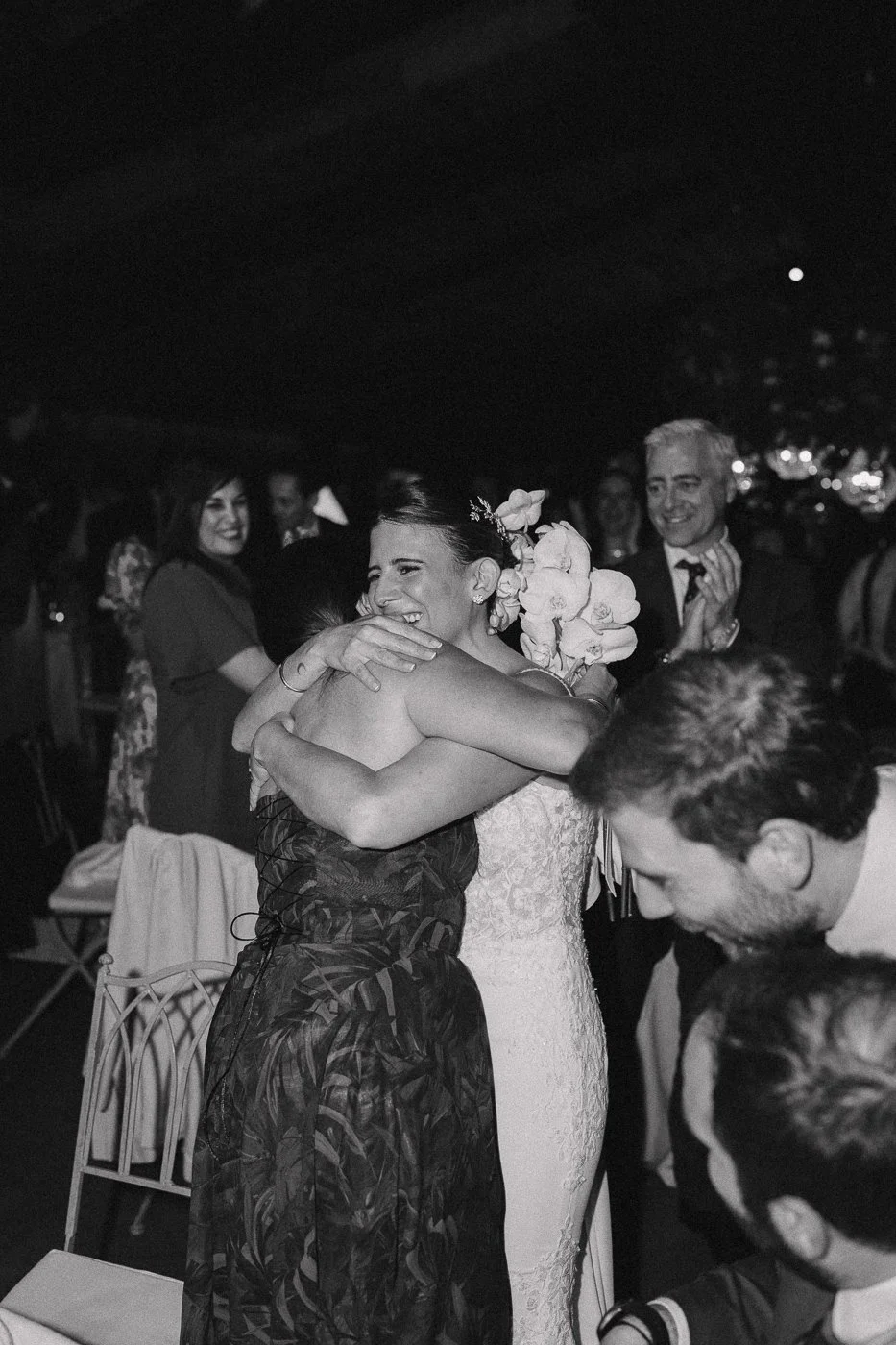 Dos mujeres abrazándose en una celebración, una de ellas con flores en el cabello y vestida de novia, otras personas en el fondo aplaudiendo y sonriendo.