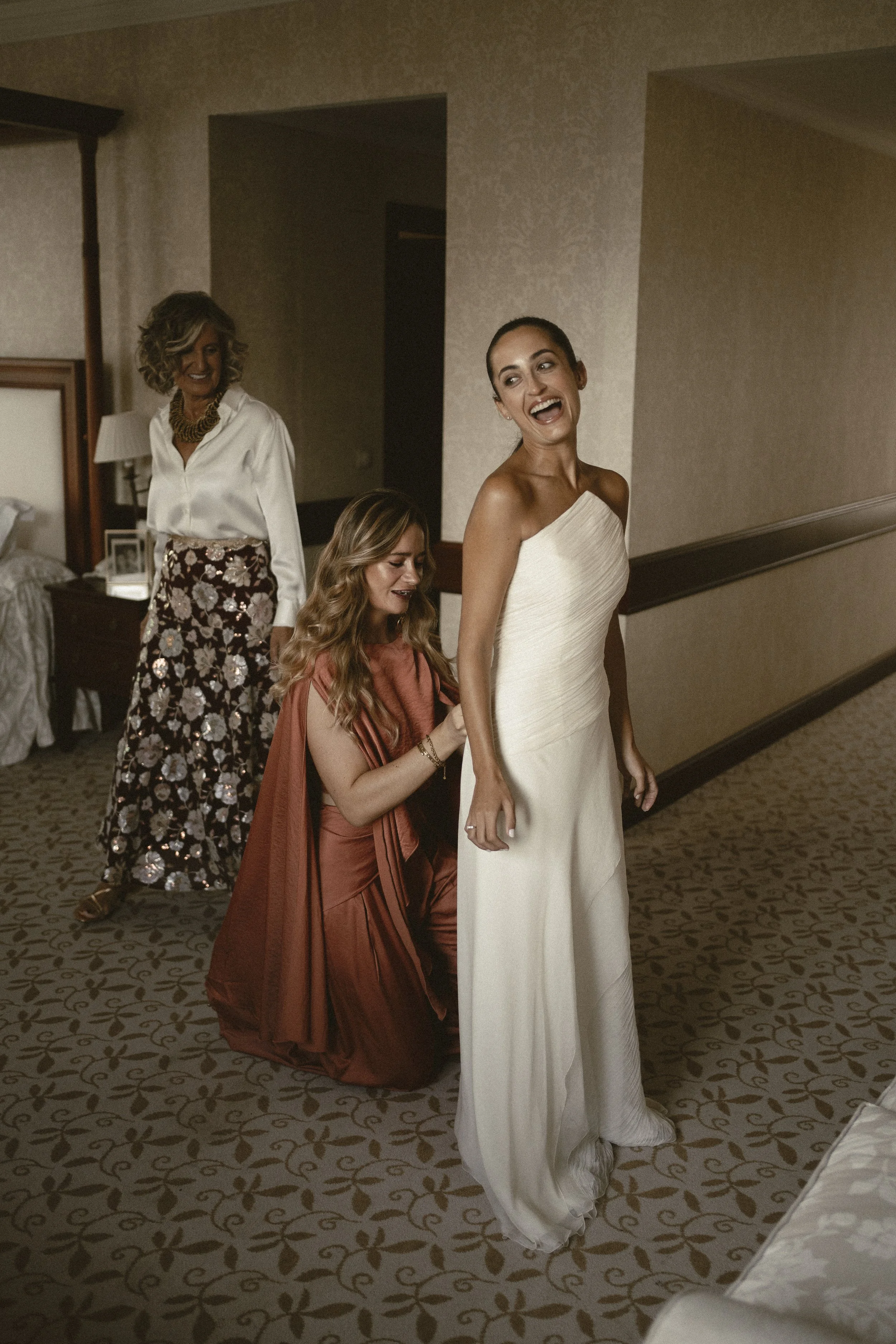 Mujer sonriendo con vestido blanco, acompañada por tres mujeres más en una habitación, preparándose posiblemente para un evento especial.