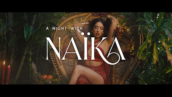 A woman in a red outfit sitting on a decorative chair surrounded by green plants, with text overlay reading 'A Night with Naika'.