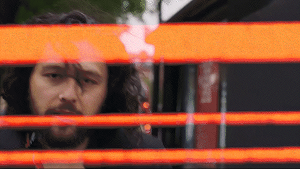 A man with long, curly hair and a beard peeks through orange construction tape, with a serious expression on his face.