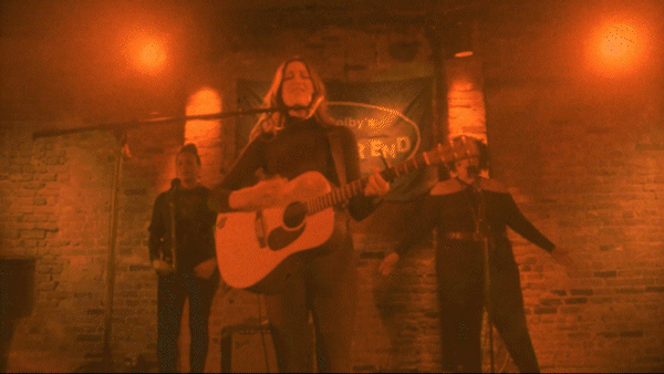 A woman singing and playing an acoustic guitar on a dimly lit stage with two backup performers behind her.
