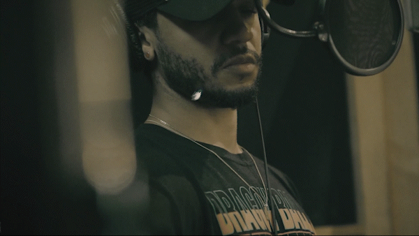 A man in a recording studio wearing headphones and a black T-shirt, singing or speaking into a microphone.