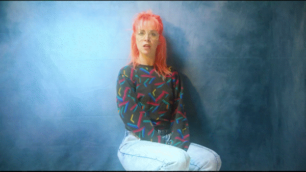 A woman with pink hair sitting against a blue wall, wearing a black shirt with colorful patterns and white pants.